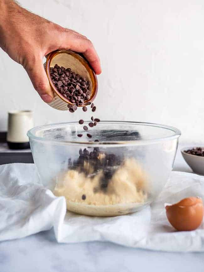 hand pouring chocolate chips into cookie dough inside a glass bowl.
